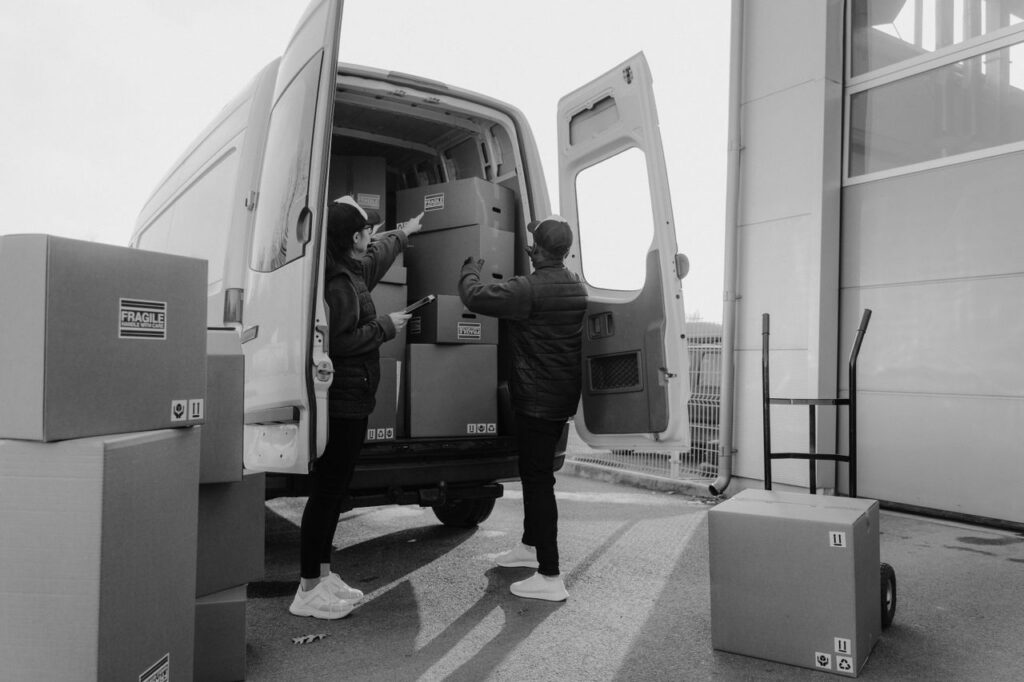 Two couriers loading cardboard boxes into a delivery van outdoors.